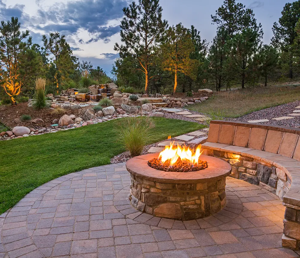 Outdoor space with paver patio, firepit with lit fire, retaining wall with sitting area, and large water feature in the background