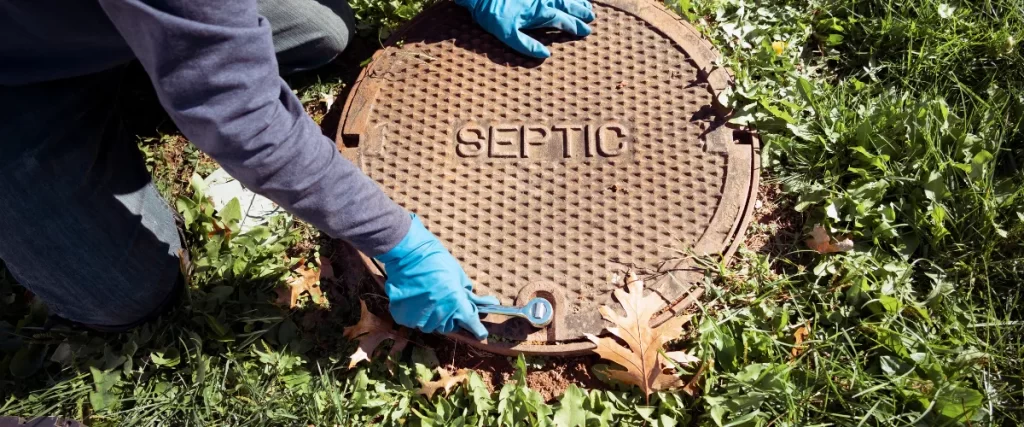 Worker wearing blue gloves opening a round septic tank lid labeled "SEPTIC" with a tool.