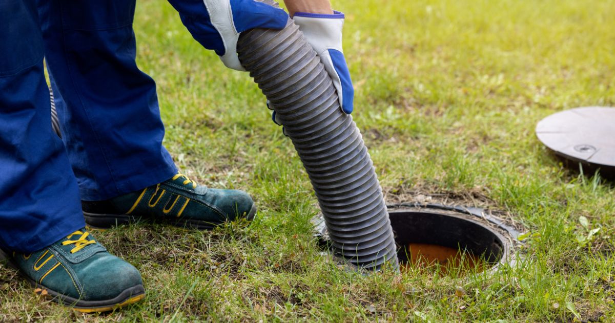 Person in blue gloves holding a large hose to pump waste from an underground septic tank.