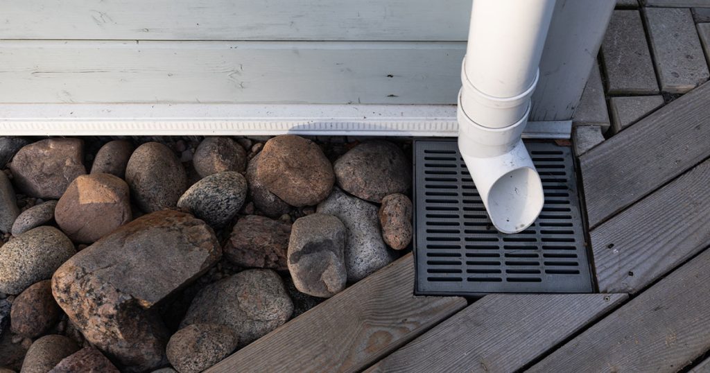 House downspout draining rainwater into ground drainage system with rocks and deck for water runoff control