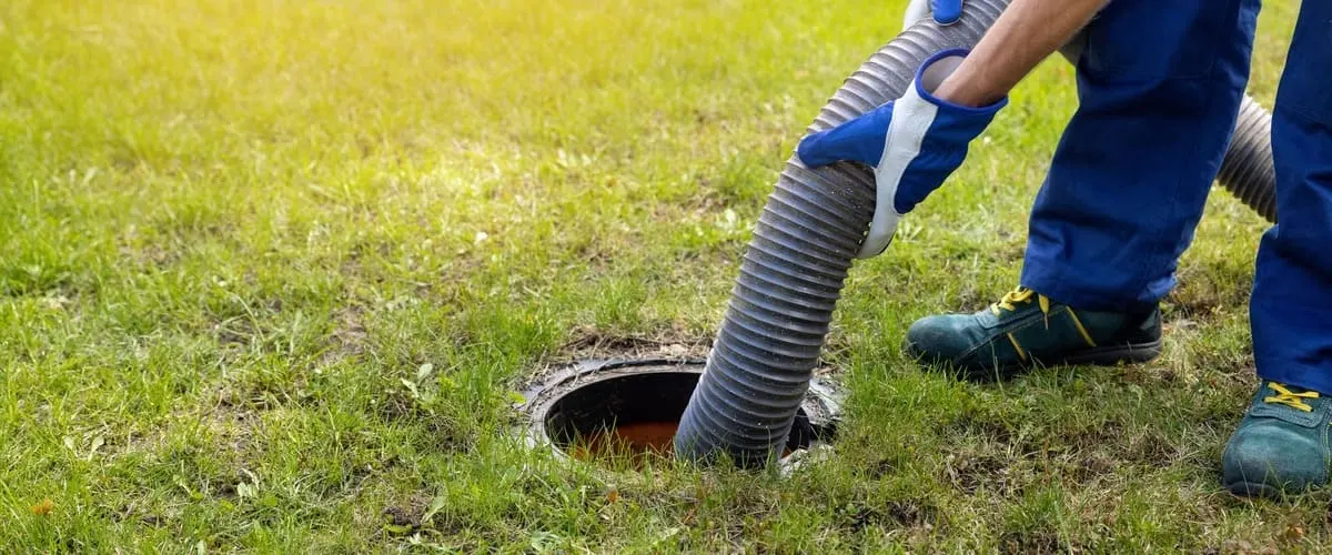 A professional technician wearing blue safety gloves holds a large corrugated vacuum hose inserted into a ground access point during routine system maintenance.