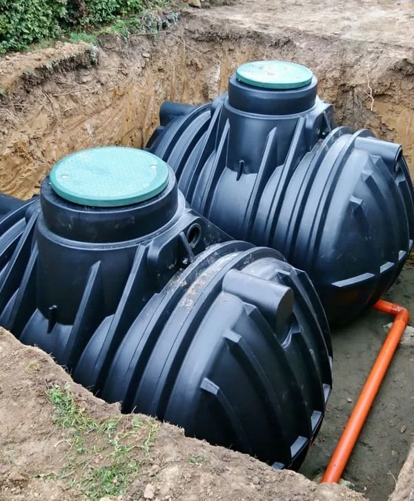 Two heavy-duty black polyethylene septic tanks connected by orange piping sitting in a deep dirt excavation pit during a new septic tank installation.