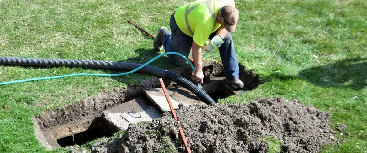 A worker in a neon yellow safety shirt kneels beside an open dirt trench to guide a cleaning hose into an exposed underground drain pipe.