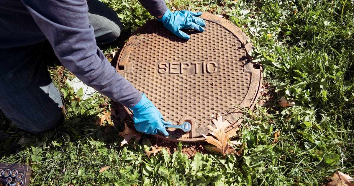 A professional service technician wearing blue protective gloves uses a wrench to unscrew the bolt on a heavy rusted metal septic tank cover labeled SEPTIC to show how septic system works during a routine inspection.