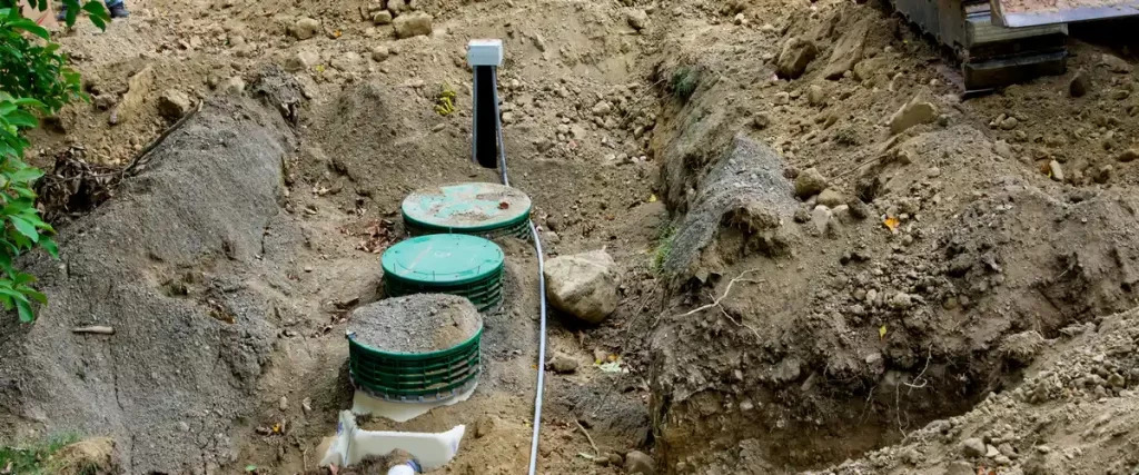 An aerial view of a deep dirt trench containing green septic system risers and connecting pipes during the construction phase.