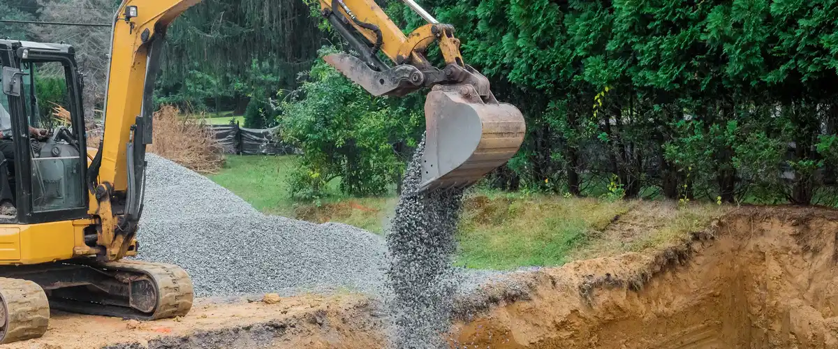 Close-up of an excavator bucket pouring grey gravel into a freshly dug trench for drainage during excavation.