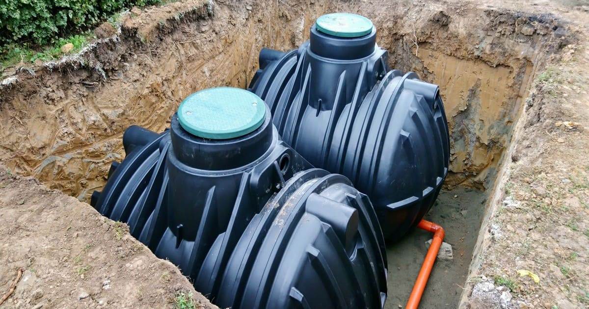 Professional septic tank installation showing two large black plastic underground holding tanks with green lids being placed in a excavated dirt pit.