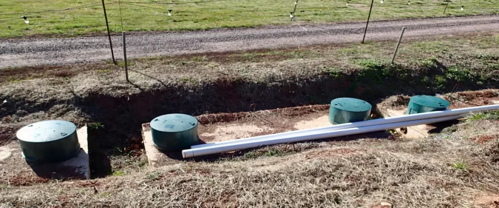 Multiple green septic system risers and white PVC pipes situated on a grassy mound area next to a gravel driveway.